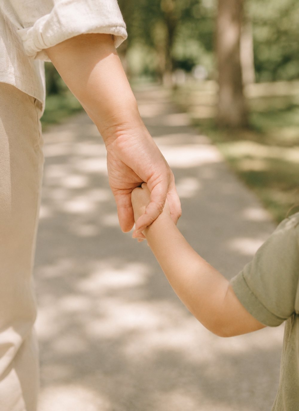 An adult holding a small child's hand on a sunlit path